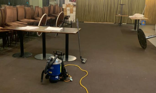 High-angle shot of a commercial cleaning in progress at a Barangaroo event venue. A blue commercial vacuum cleaner with a long yellow cord sits on a large dark carpeted area, surrounded by stacked chairs and event tables.